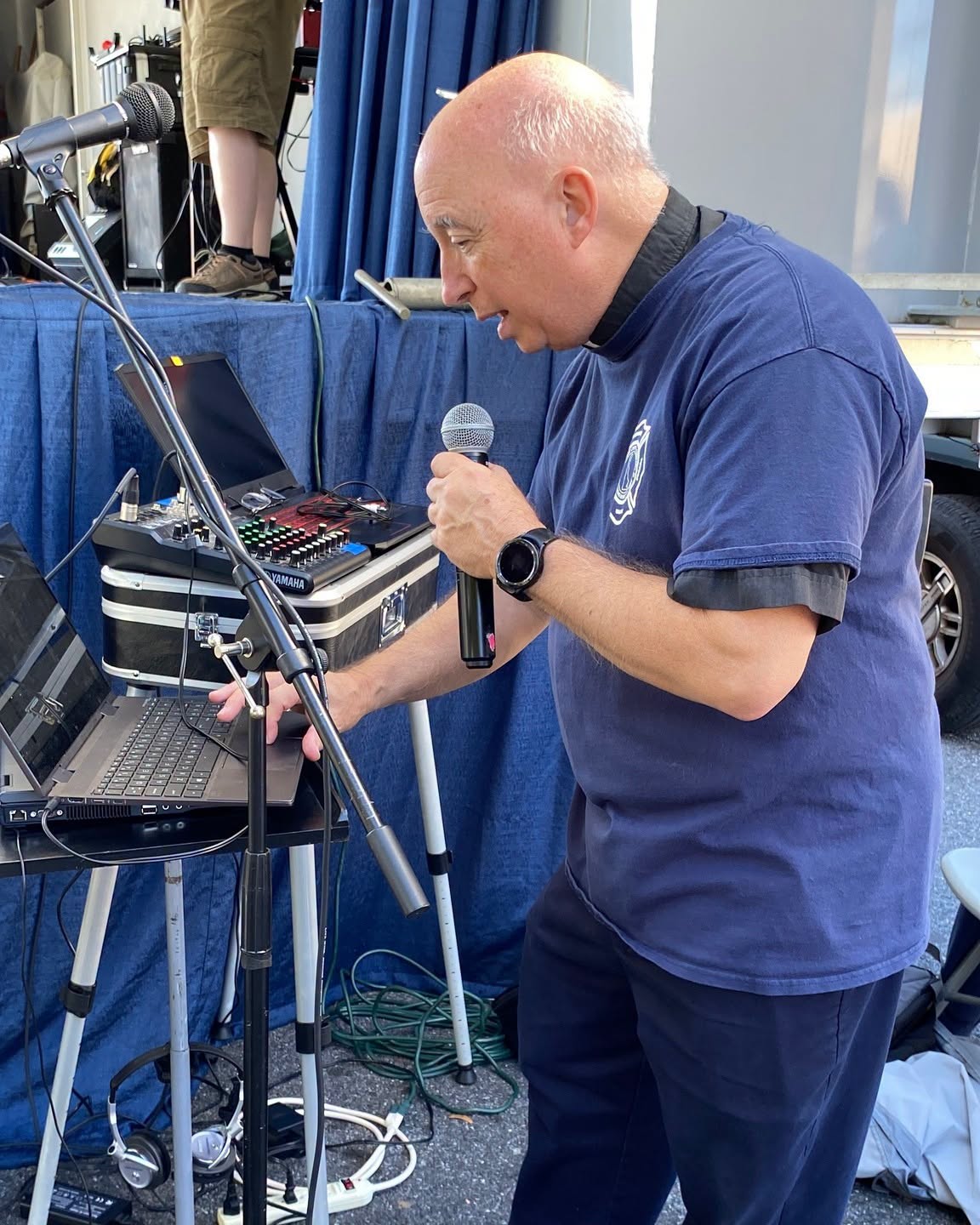 A host speaking into a microphone beside an audio mixing board