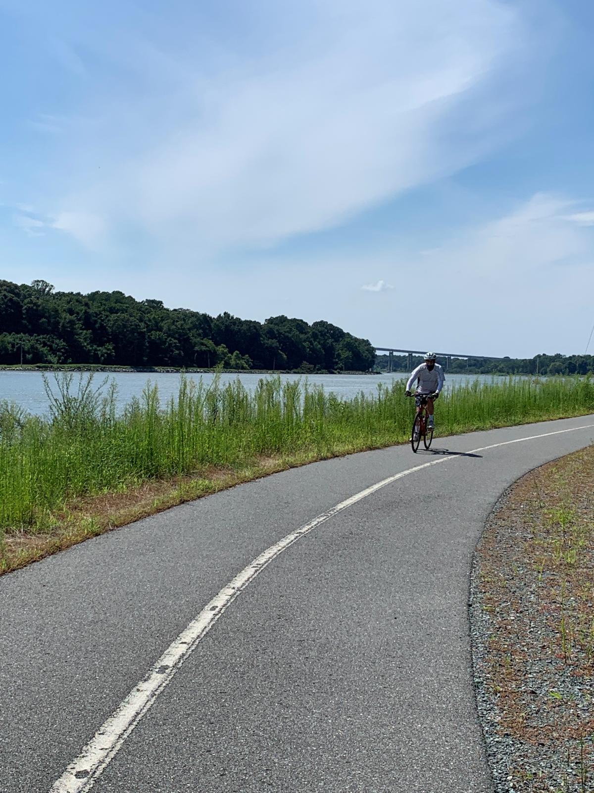 A cyclist on the paved Michael N. Castle Trail beside the canal, with grasses, trees and a bridge in the distance