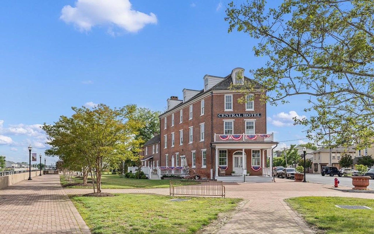 The Central Hotel, a three-story red-brick building with a covered porch, white columns, a balcony and patriotic bunting, beside a brick-paved canal-front promenade in Delaware City