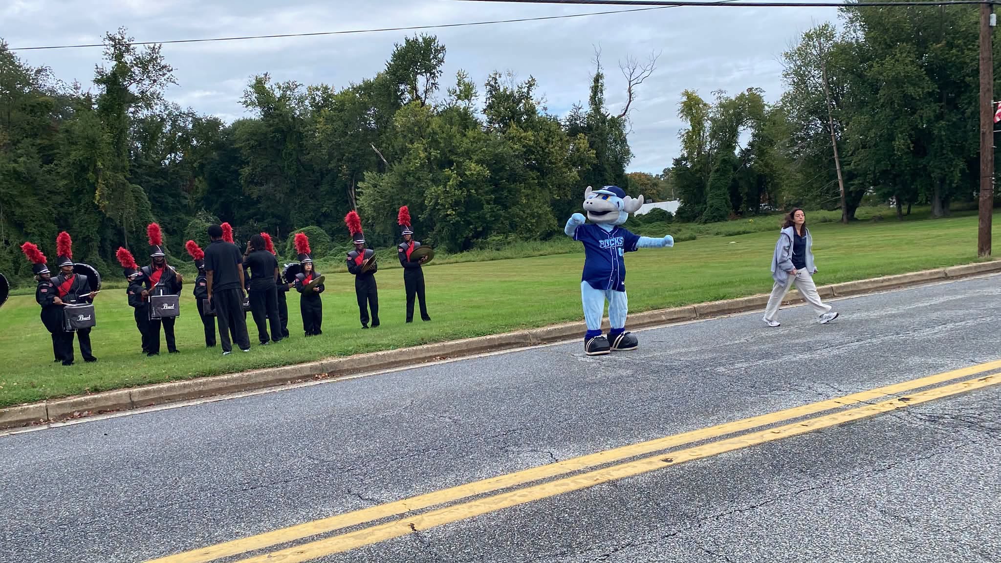 A blue costumed mascot leading a marching band along the parade route