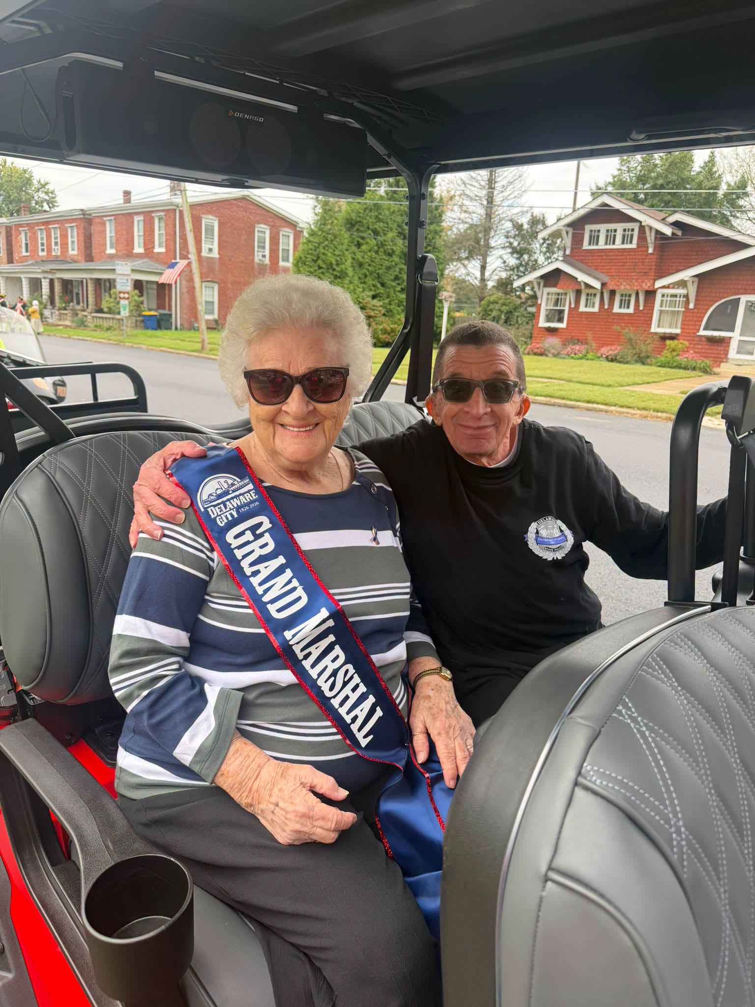 A parade Grand Marshal wearing a sash beside a golf cart