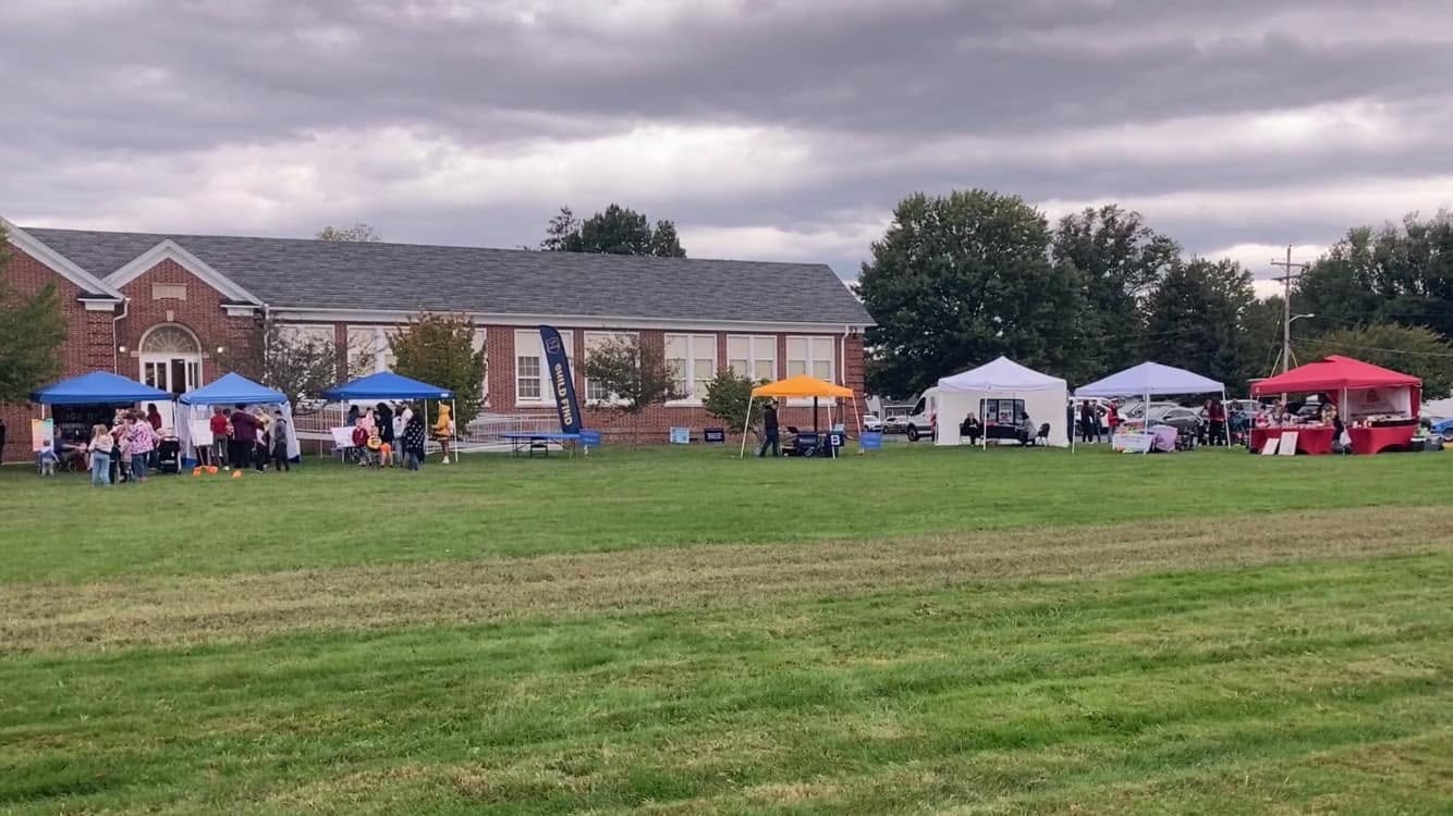 Rows of festival vendor tents spread across a grass field beside a brick building