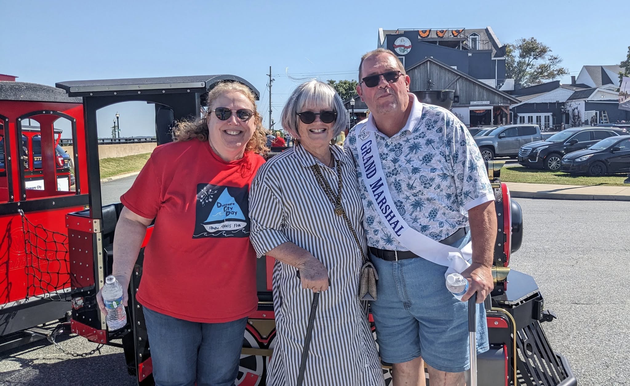 Three adults posing next to the festival train, one wearing a Grand Marshal sash